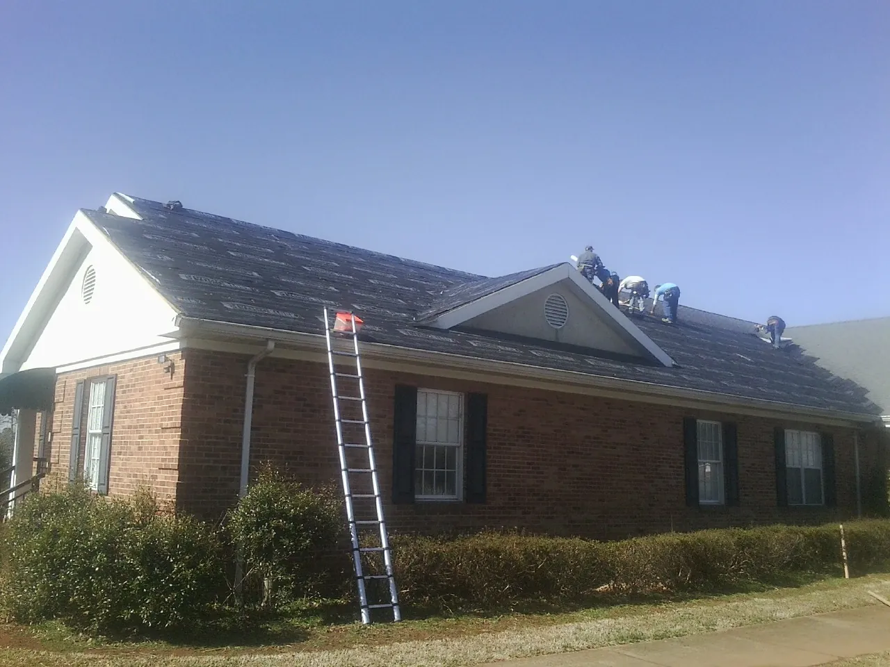 Crew tearing shingles off a brick ranch roof with a ladder set — Emergency Roof Repair work by our crew in Pledger