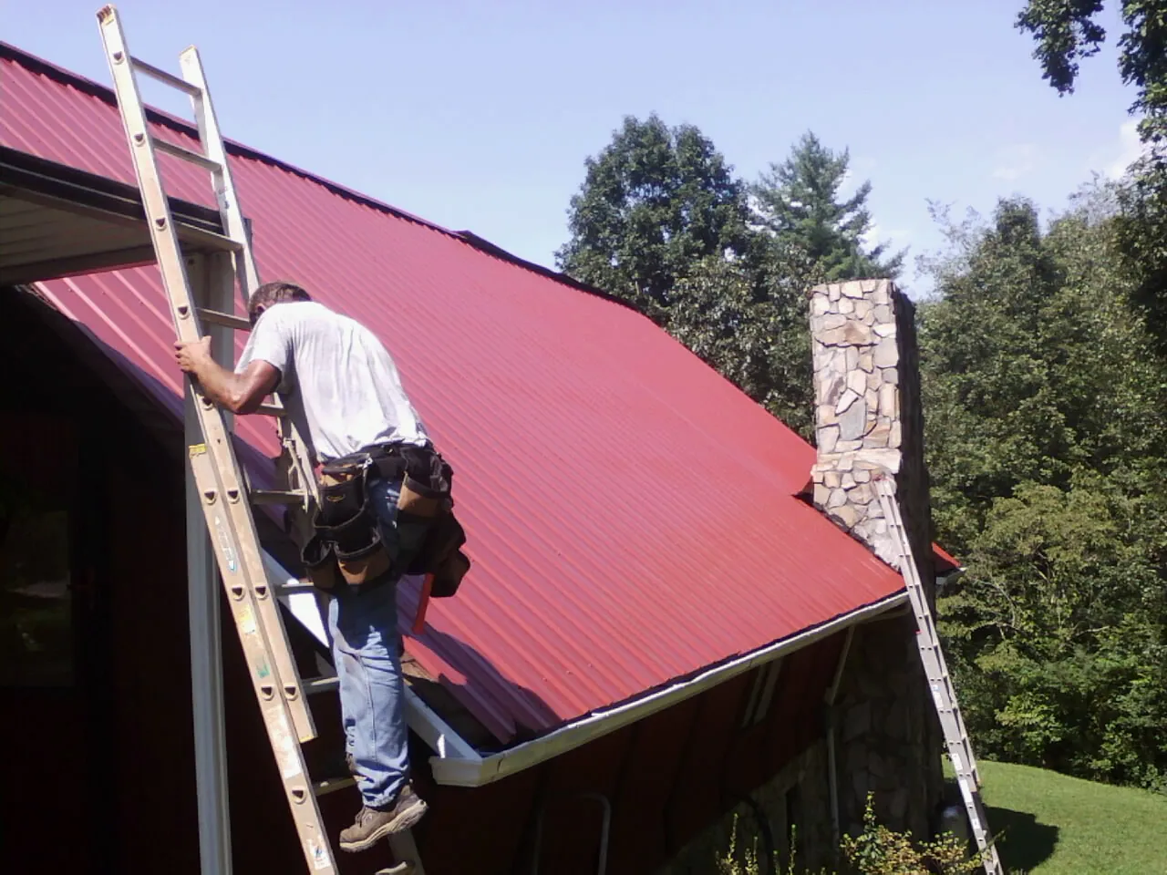 Roofer on a ladder beside a red metal roof and stone chimney — Soffit & Fascia Repair by Roofing Brothers in Pledger